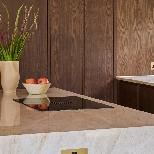 Wooden kitchen shelves with plates, bowls, and vintage books. A bowl of apples rests on the middle shelf. Below, sprigs of dried herbs hang above a marble countertop with a dish towel on the side