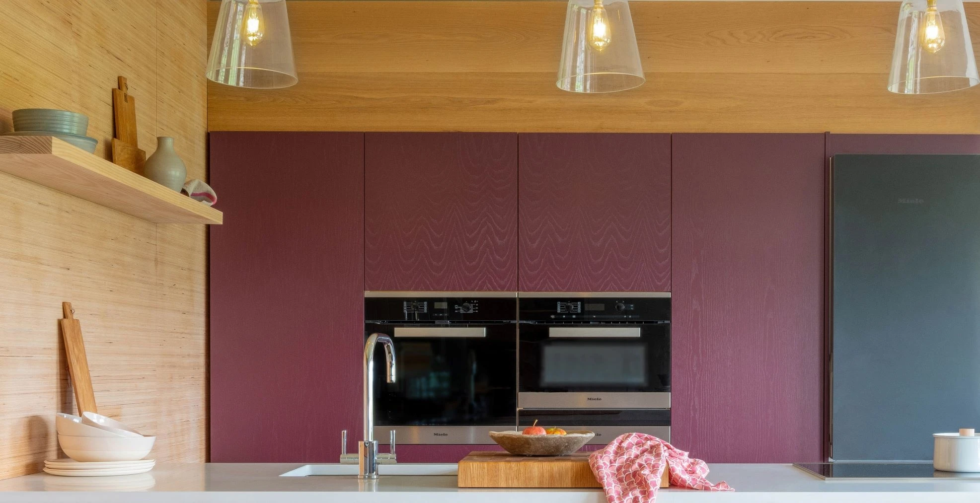 Modern kitchen with purple cabinetry, double ovens, and a sleek gray refrigerator. A countertop features a wooden cutting board, a bowl of fruit, and a patterned cloth. Transparent pendant lights hang from the wooden ceiling. Minimalist decor on shelves.