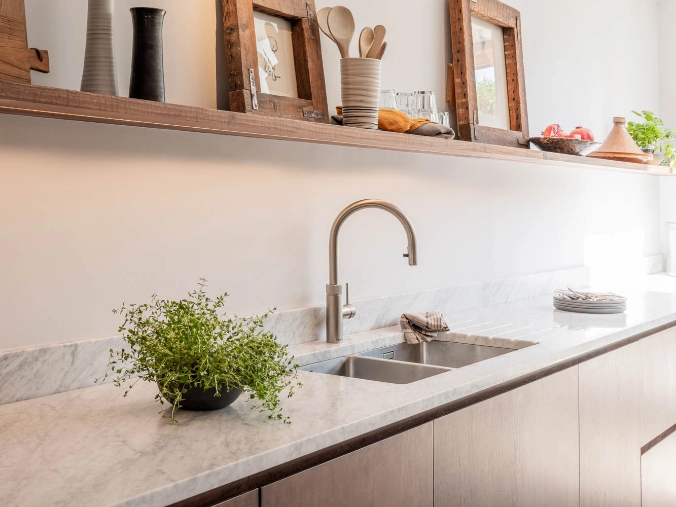 A photograph of a modern kitchen featuring a marble countertop with a stainless steel undermount sink, a sleek arched faucet, and a wooden open shelf displaying rustic decor and kitchenware.