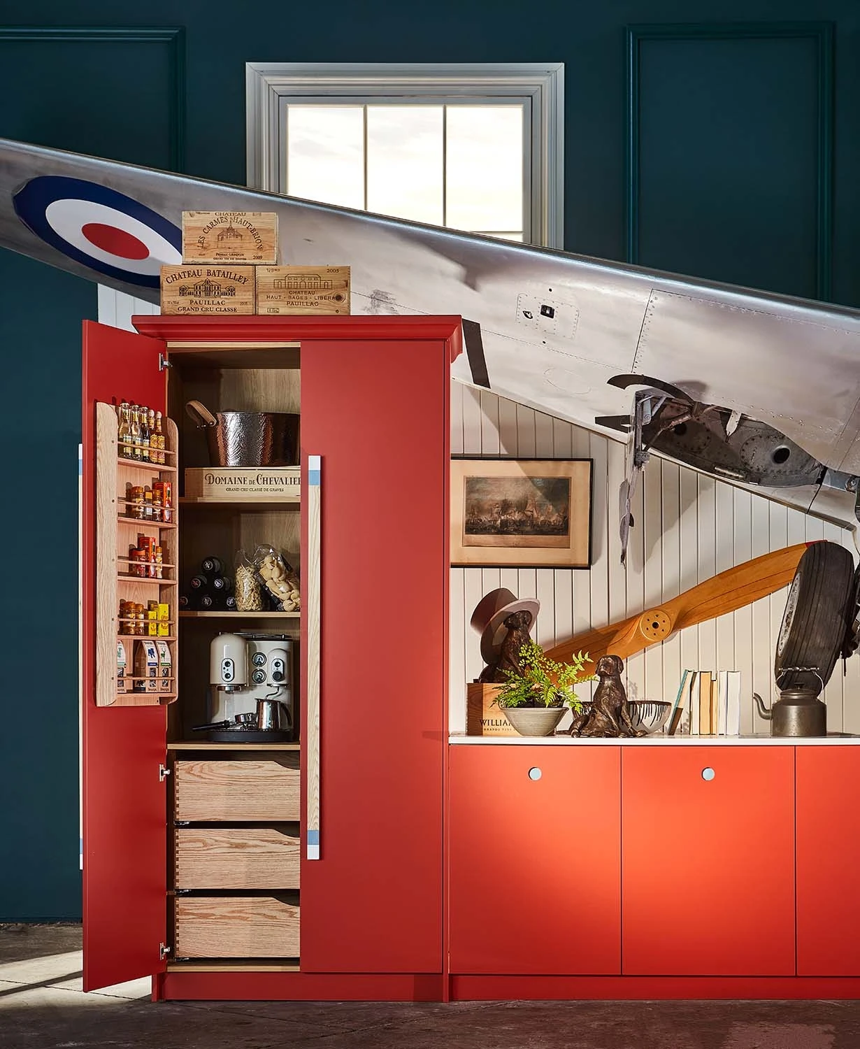 A unique kitchen setup featuring a bright red pantry with various items, against a backdrop of an airplane wing extending into the room. The countertop displays decorative items, including a propeller piece and a vintage figurine