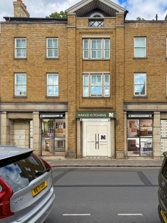 Street view of the Naked Kitchens showroom in a three-story yellow brick building. The central entrance features a green sign and white doors labeled Coming Soon. Flanking windows for Naked Designs and Naked Studio display various interior kitchen and furniture styles.