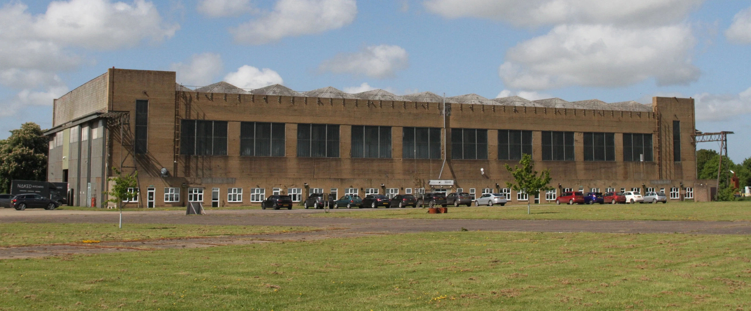 A large, rectangular industrial building under a partly cloudy blue sky. The building has multiple windows and parked cars in front. The grass field in the foreground is expansive and green.