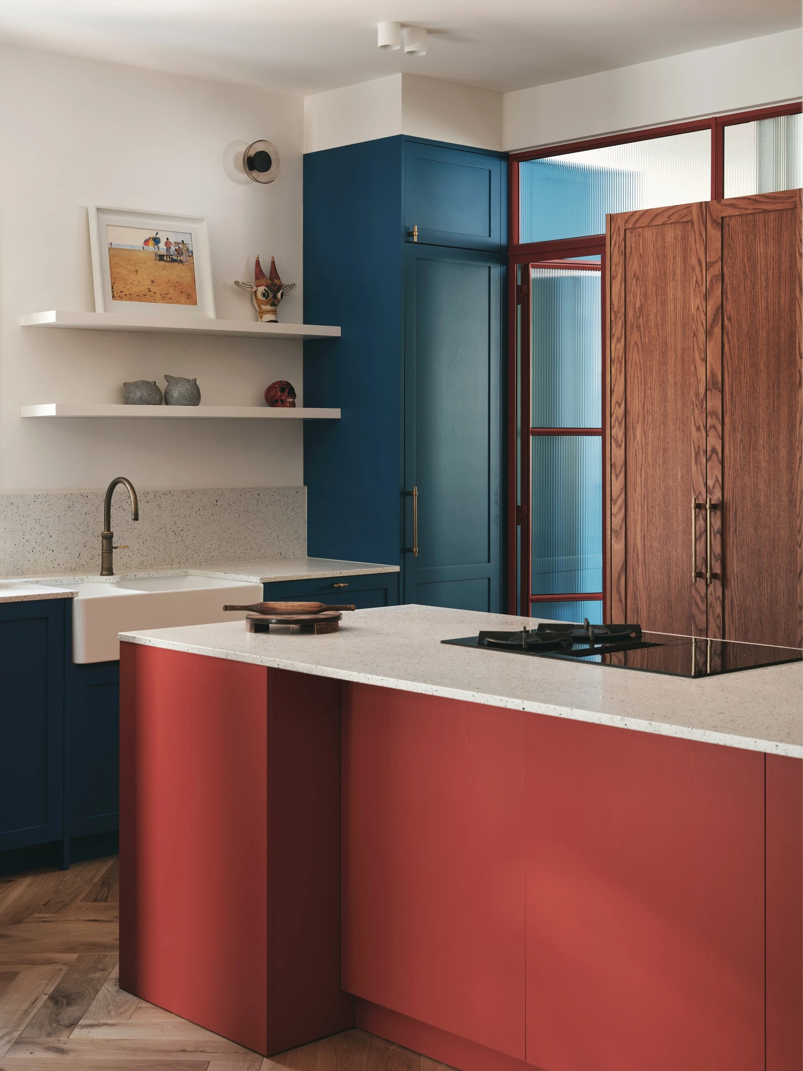Modern kitchen with red and blue cabinetry, a white countertop island with a built-in stovetop, and an undermount sink. Shelves hold decor, and a large wooden door is in the background. Light filters in softly, highlighting the sleek design.