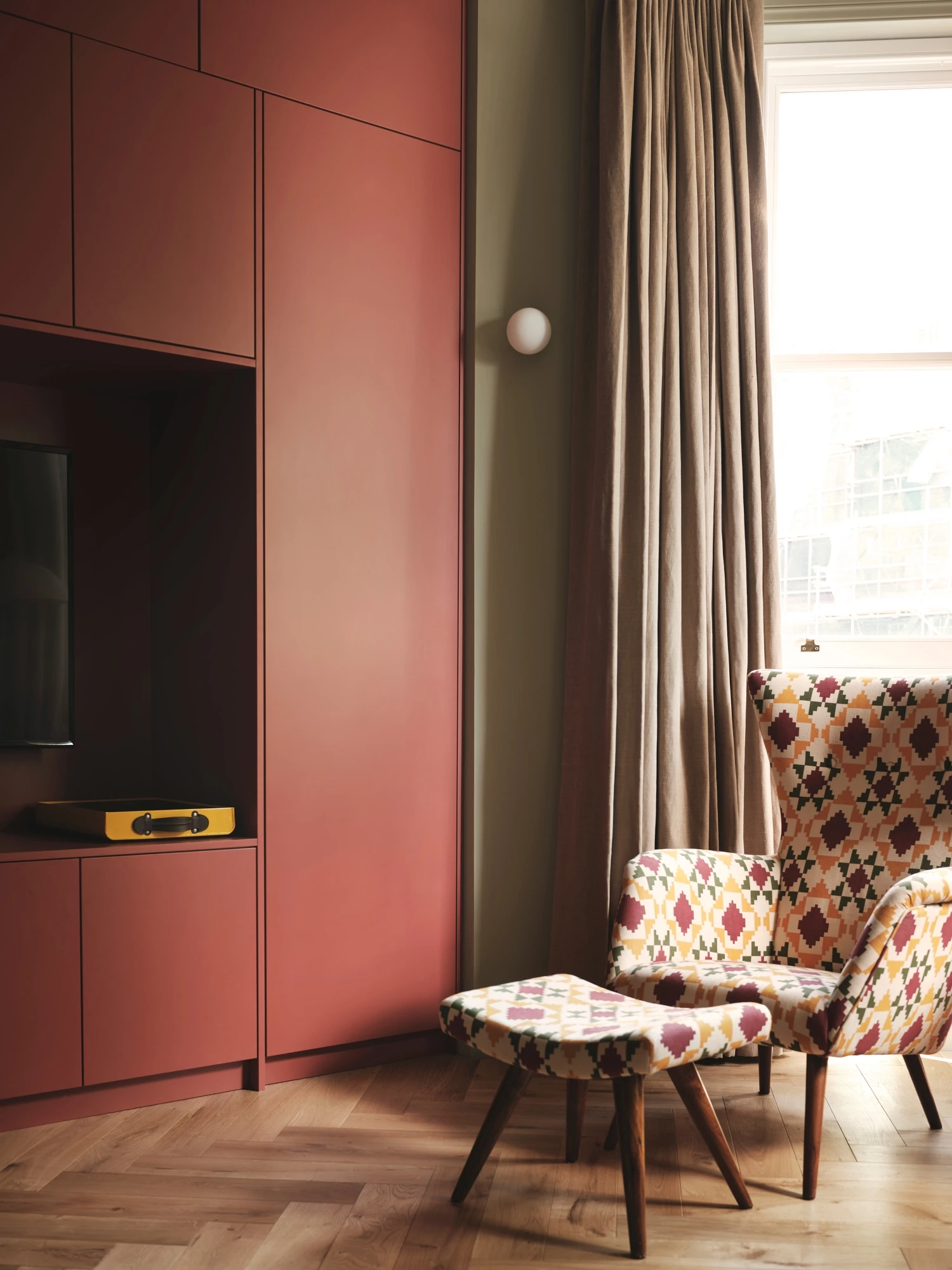 A cozy living room corner features a patterned wingback chair with matching ottoman, set against reddish-brown cabinetry. A wall-mounted TV and light tan curtains add to the room's inviting atmosphere with natural light streaming in.