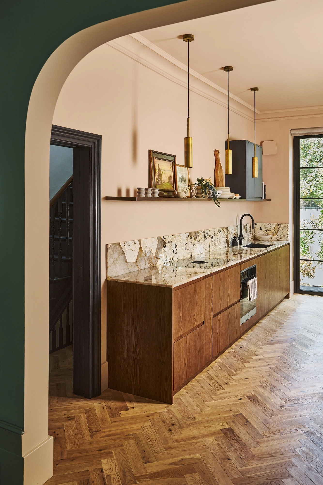 Modern kitchen with a marble hexagon-patterned splashback, wooden cupboards, black sink and cooker, gold pendant lights, and an open shelf holding crockery, chopping boards, framed art, and a potted plant.