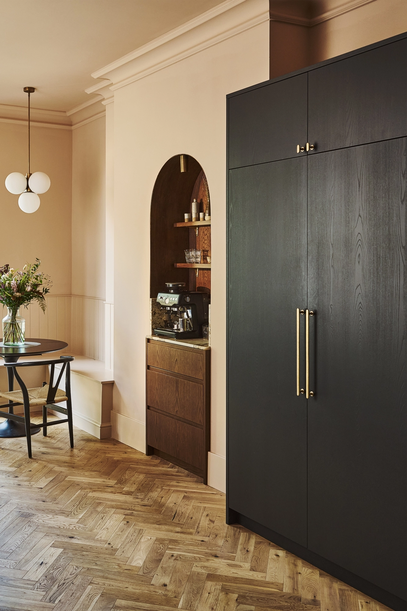 A modern kitchen with wooden cabinets, marble backsplash, a built-in oven, cookware on the stove, open shelves holding plates, bowls, framed art, and decorative items, lit by two gold pendant lights.