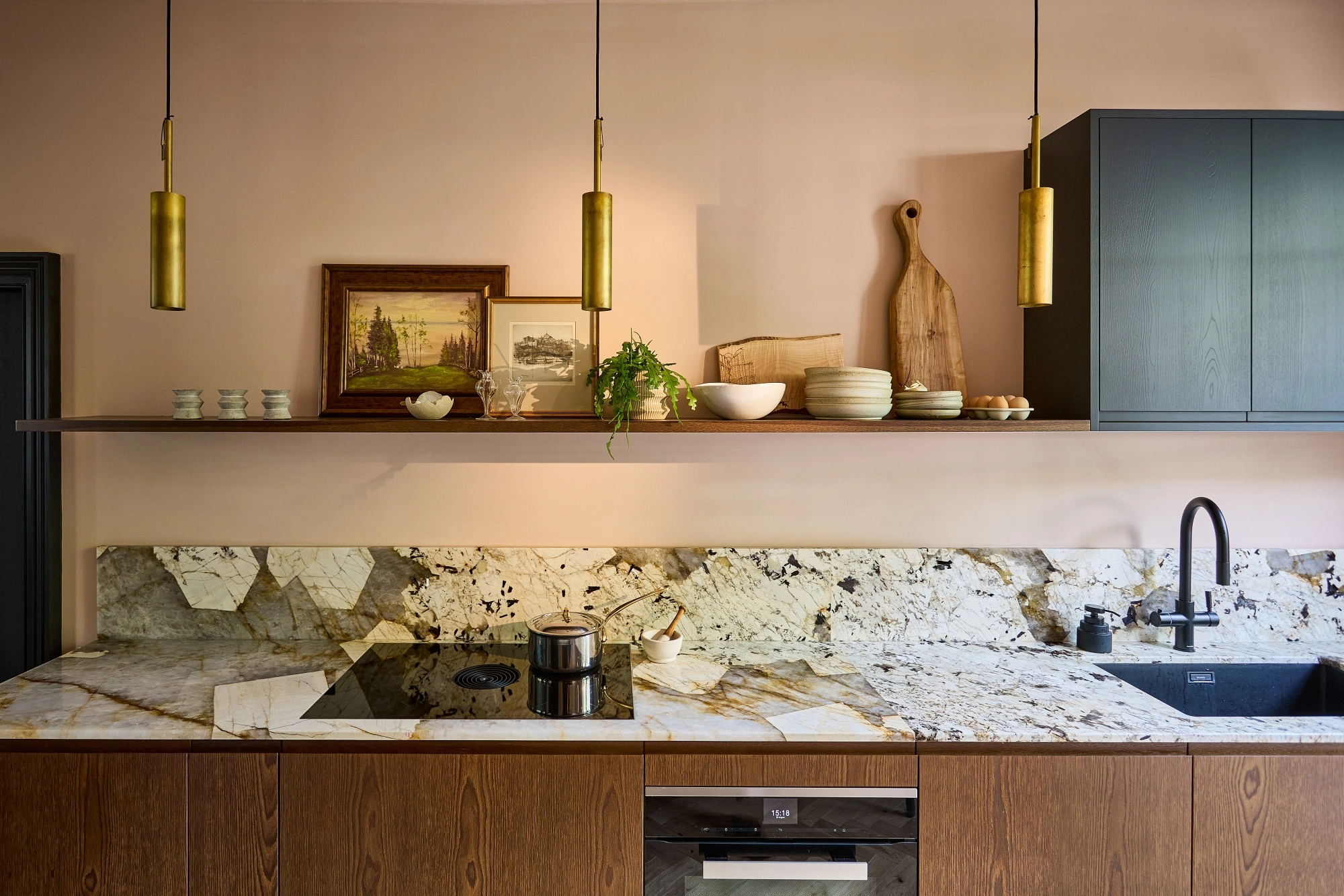 Modern kitchen with a marble hexagon-patterned splashback, wooden cupboards, black sink and cooker, gold pendant lights, and an open shelf holding crockery, chopping boards, framed art, and a potted plant.