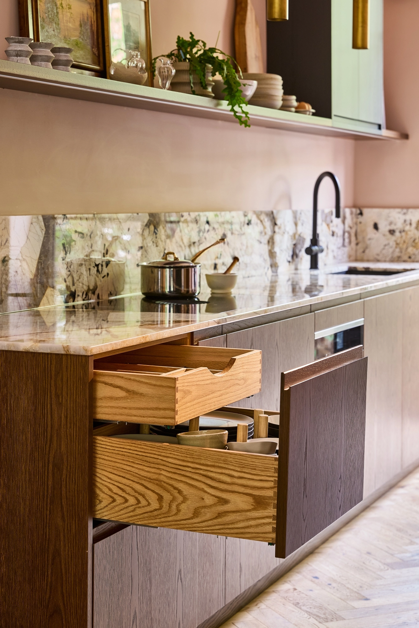 Close-up of a modern kitchen with wooden cabinets, marble backsplash, a built-in oven, cookware on the stove, open shelves holding plates, bowls, framed art, and decorative items, lit by two gold pendant lights.