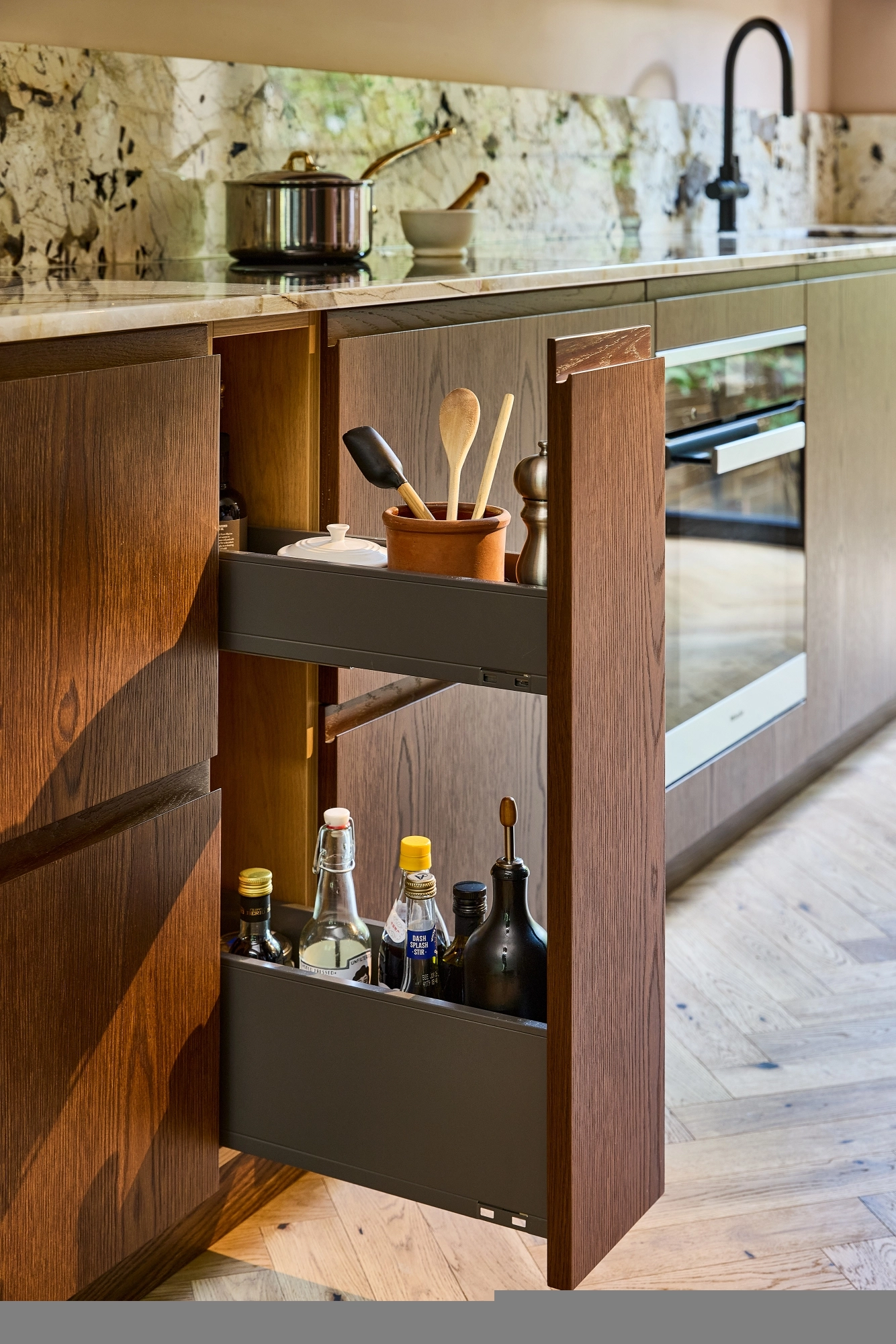 A modern kitchen features a pull-out wooden cabinet with shelves holding utensils, spices, and bottles of oil and vinegar next to an oven and sink.