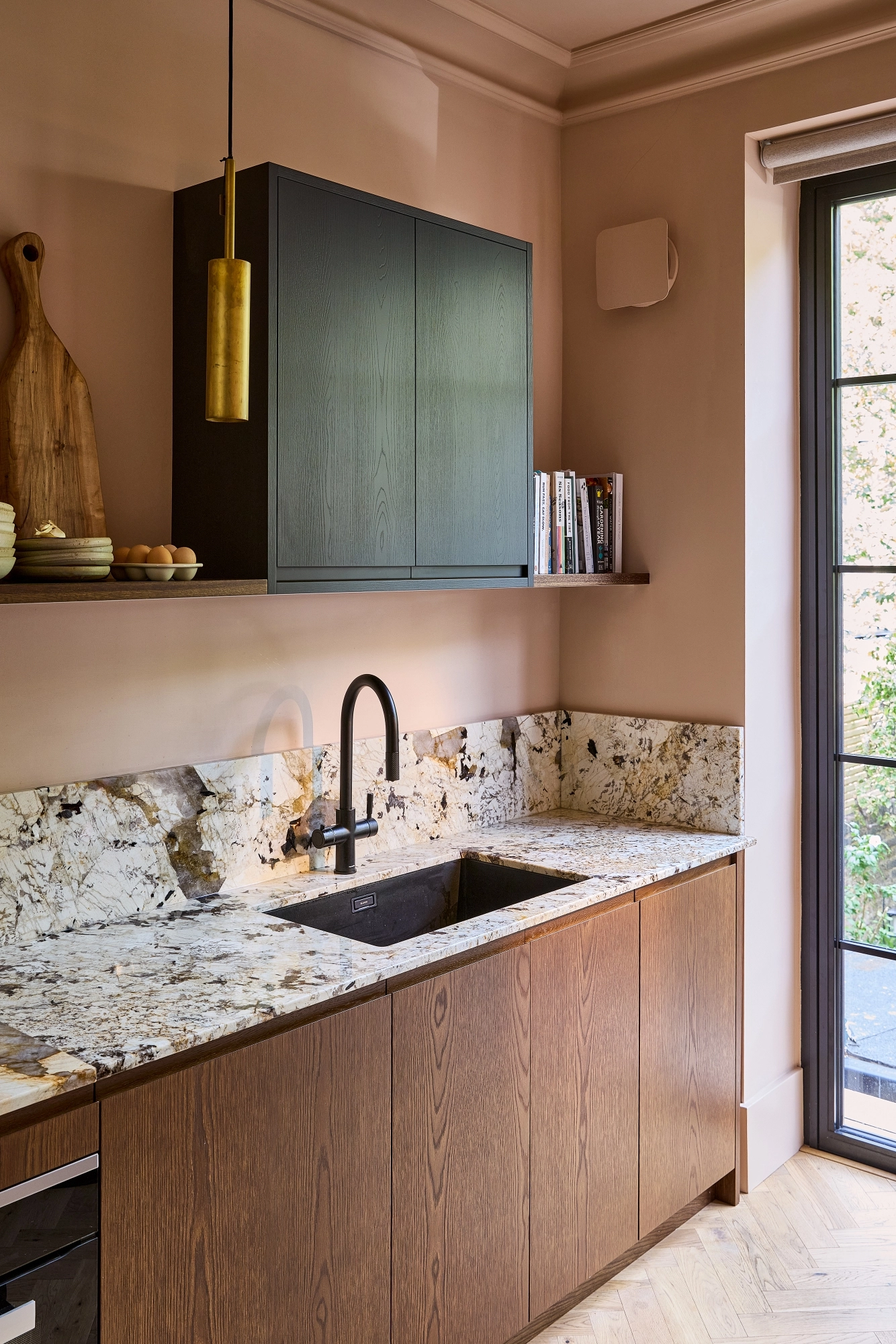 Modern kitchen with dark wood cabinets, marble countertop and backsplash, black sink and faucet, floating shelf with dishes and cookbooks, gold pendant light, and large window letting in natural light.