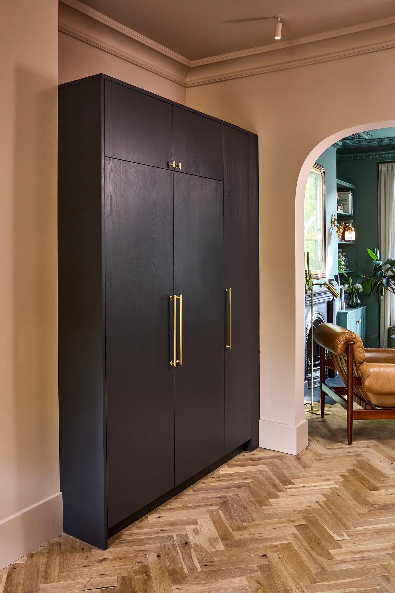 A sleek, dark wood wardrobe with gold handles stands against a beige wall near an arched doorway; parquet wood flooring and a cozy, stylish living space are visible in the background.
