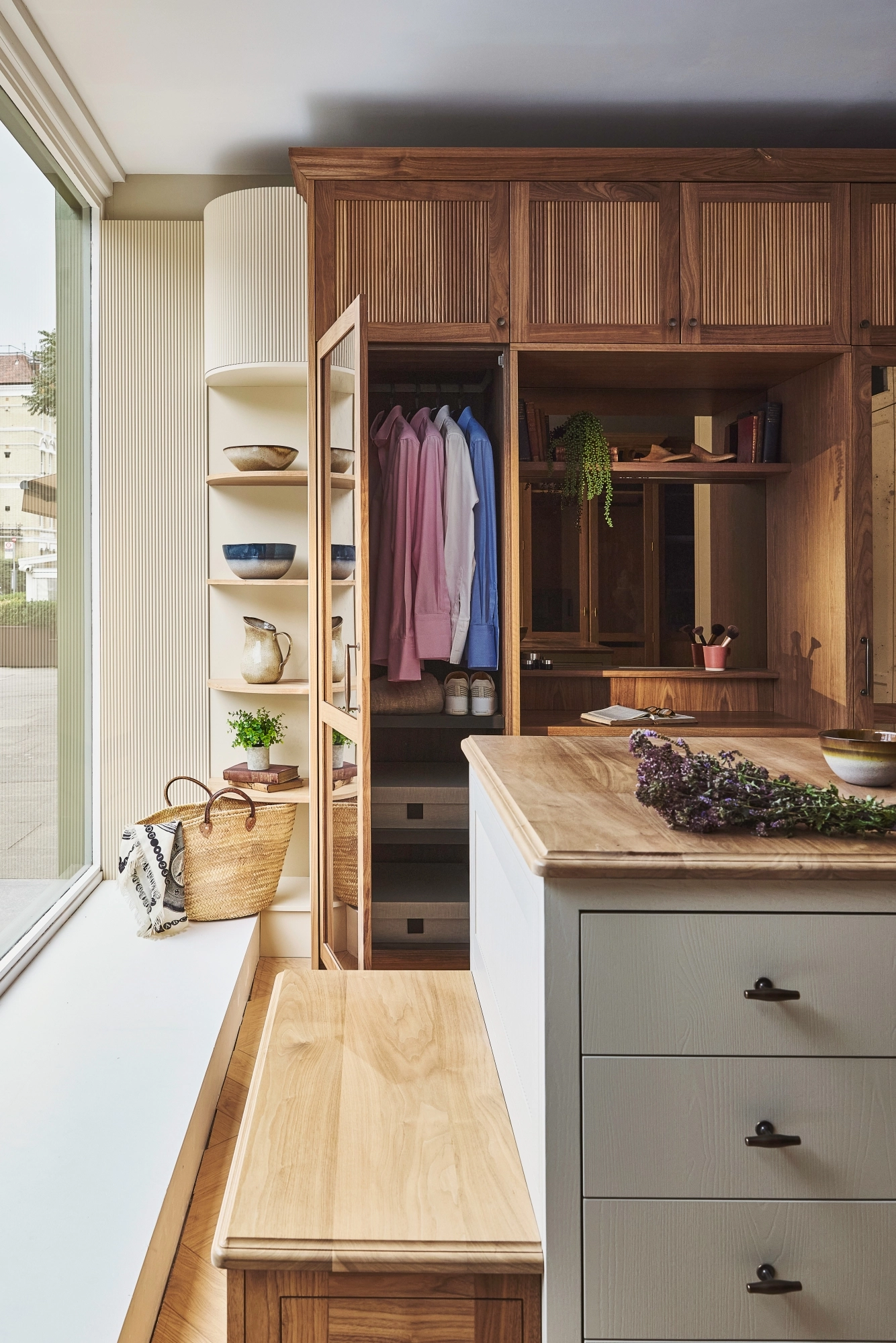 A cozy, sunlit room with wooden cabinetry, open shelves displaying bowls, a bench with a woven basket, and an island with drawers. Clothes hang in the open wardrobe, and dried flowers rest on the island.
