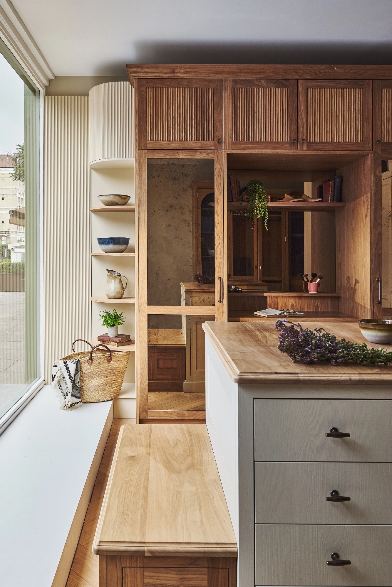 A cozy, sunlit room with wooden cabinetry, open shelves displaying bowls, a bench with a woven basket, and an island with drawers.