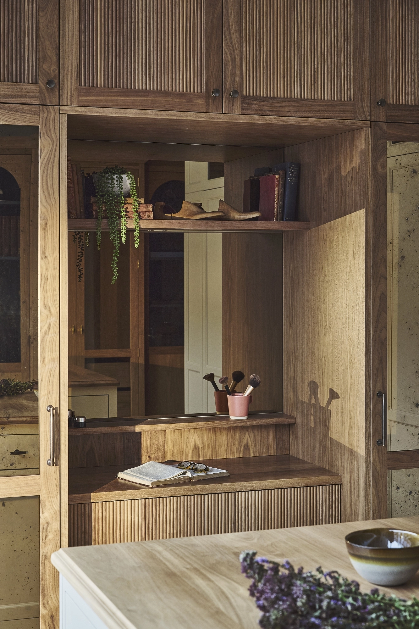 A cozy wooden vanity with shelves, books, plants, and makeup brushes sits next to a large mirror. A checkered ottoman is in front, and sunlight streams in from a window, creating a warm and inviting atmosphere.