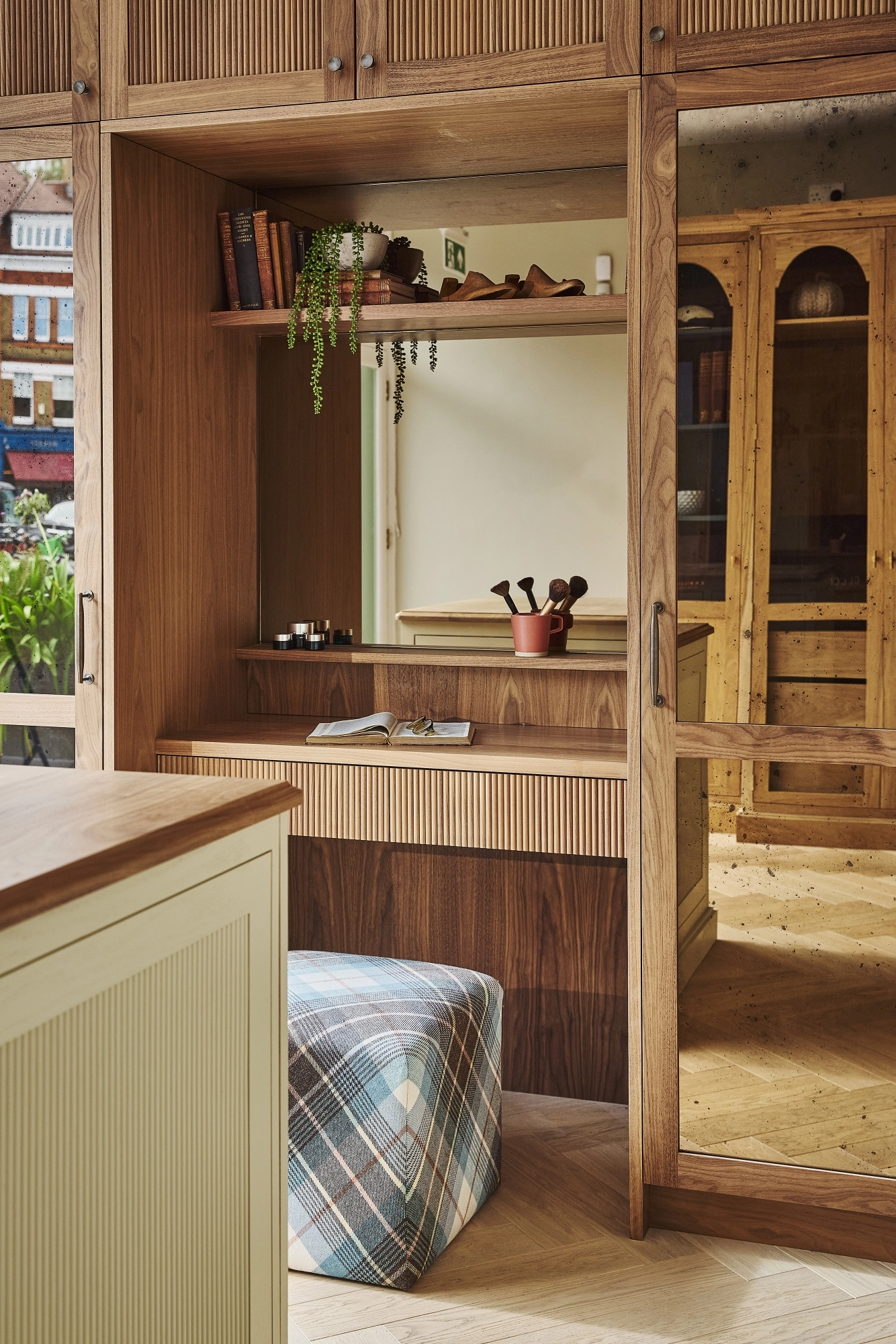A cozy wooden vanity with shelves, books, plants, and makeup brushes sits next to a large mirror. A checkered ottoman is in front, and sunlight streams in from a window, creating a warm and inviting atmosphere.