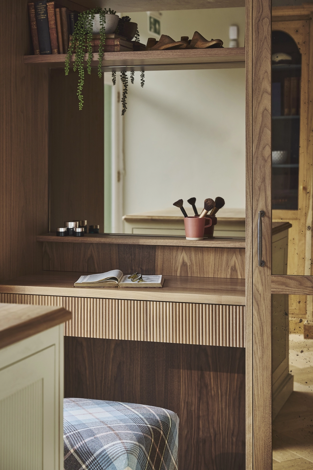 A clsoe-up of a cozy wooden vanity with shelves, books, plants, and makeup brushes sits next to a large mirror. A checkered ottoman is in front, and sunlight streams in from a window, creating a warm and inviting atmosphere.