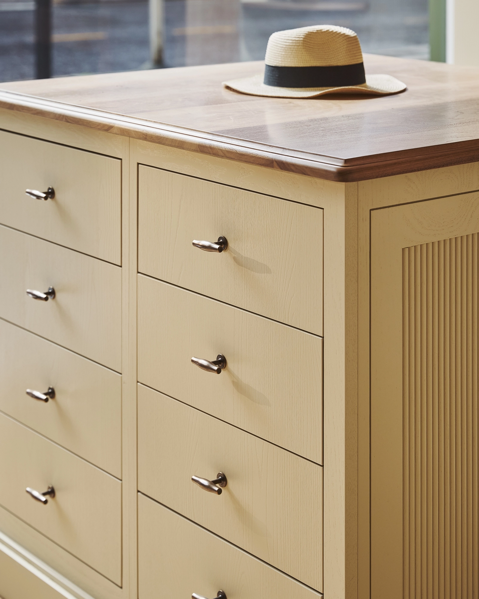 A close-up of a beige wooden cabinet with multiple drawers stands by a window in a sunlit room. A hat rests on its wooden countertop. The background shows a street scene and shops outside.