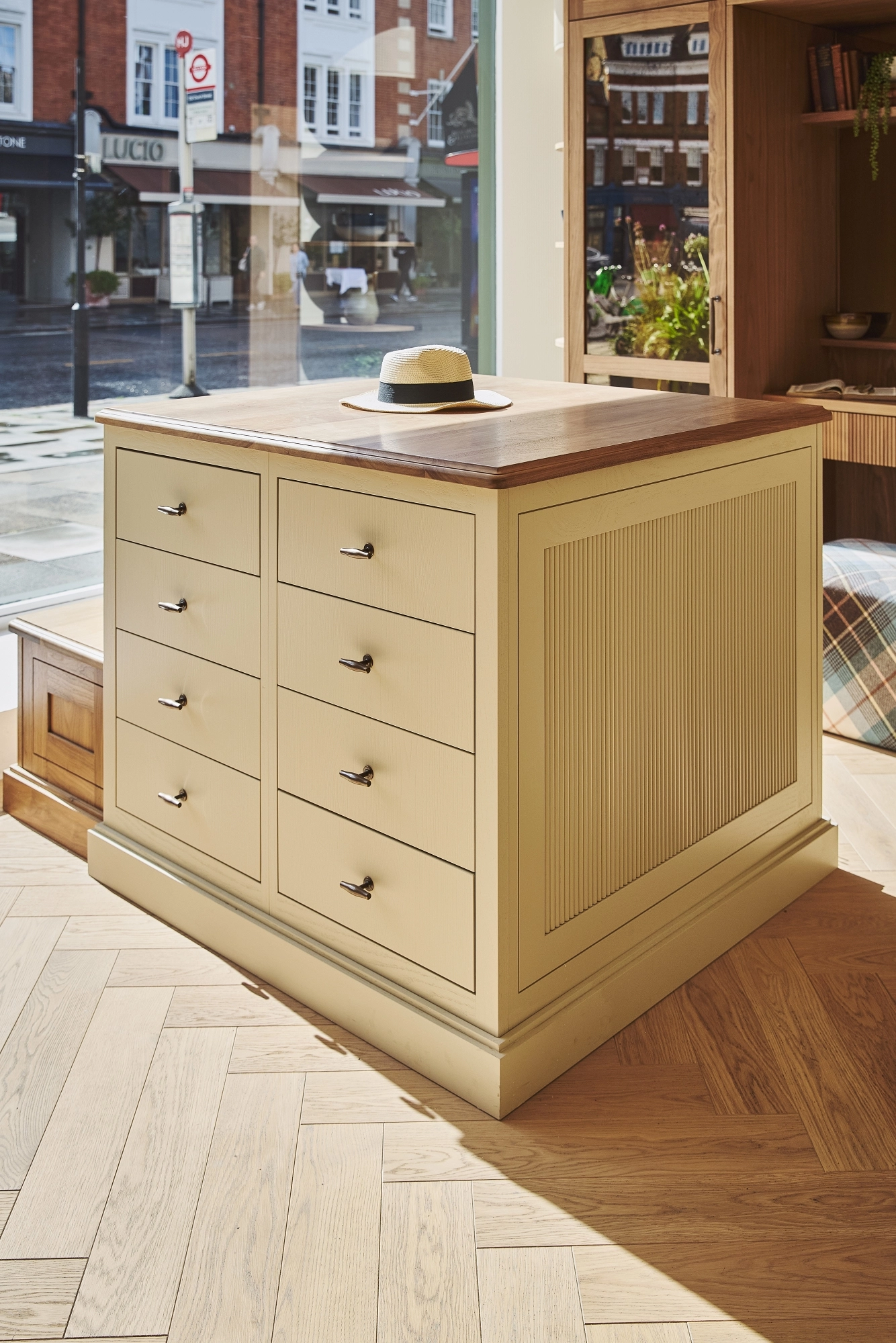 A beige wooden cabinet with multiple drawers stands by a window in a sunlit room. A hat rests on its wooden countertop. The background shows a street scene and shops outside.