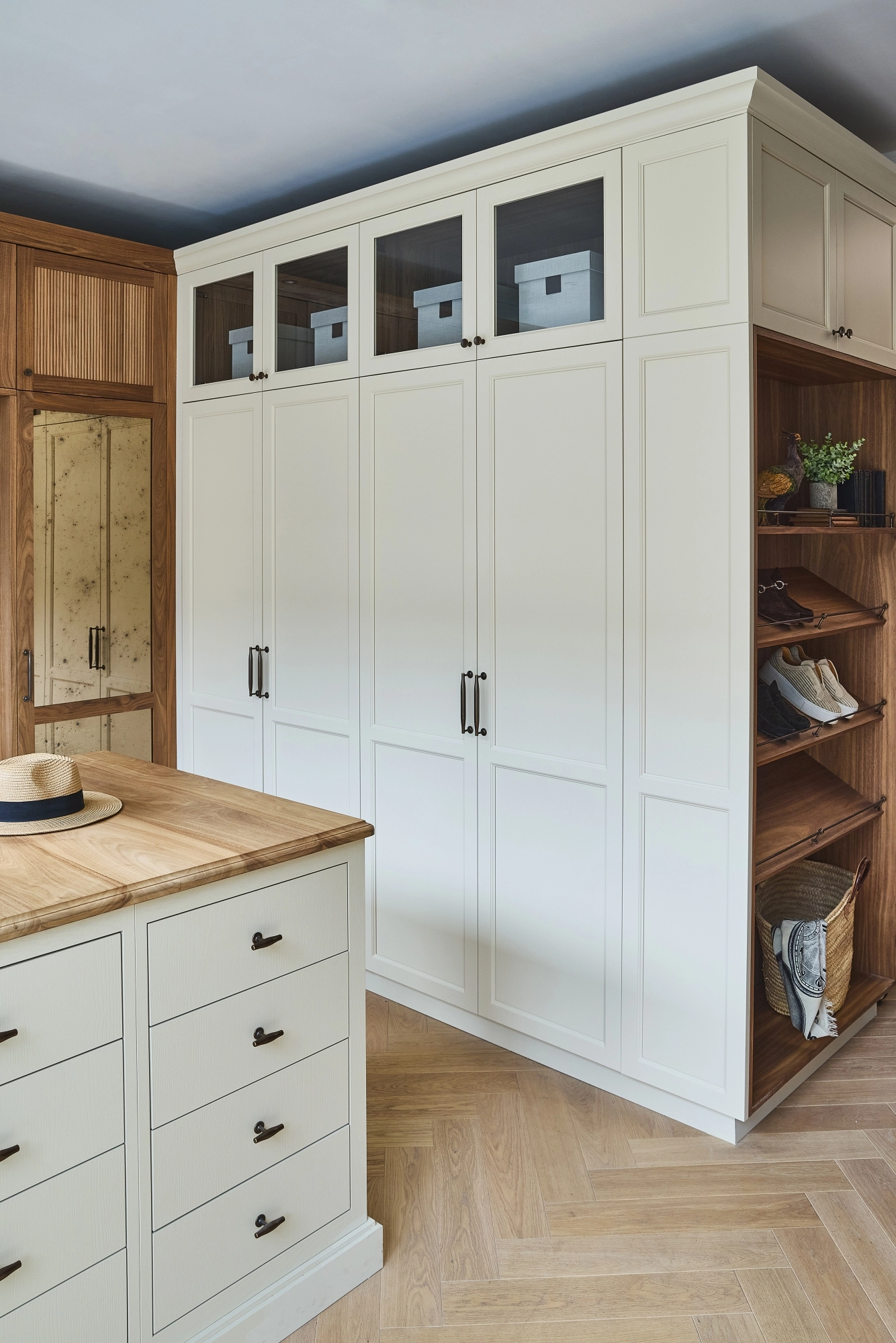 A modern walk-in closet with white cabinets, glass-paneled upper doors, wooden open shelves displaying shoes and baskets, a mirrored door, and a light wood island with drawers and a hat on top.