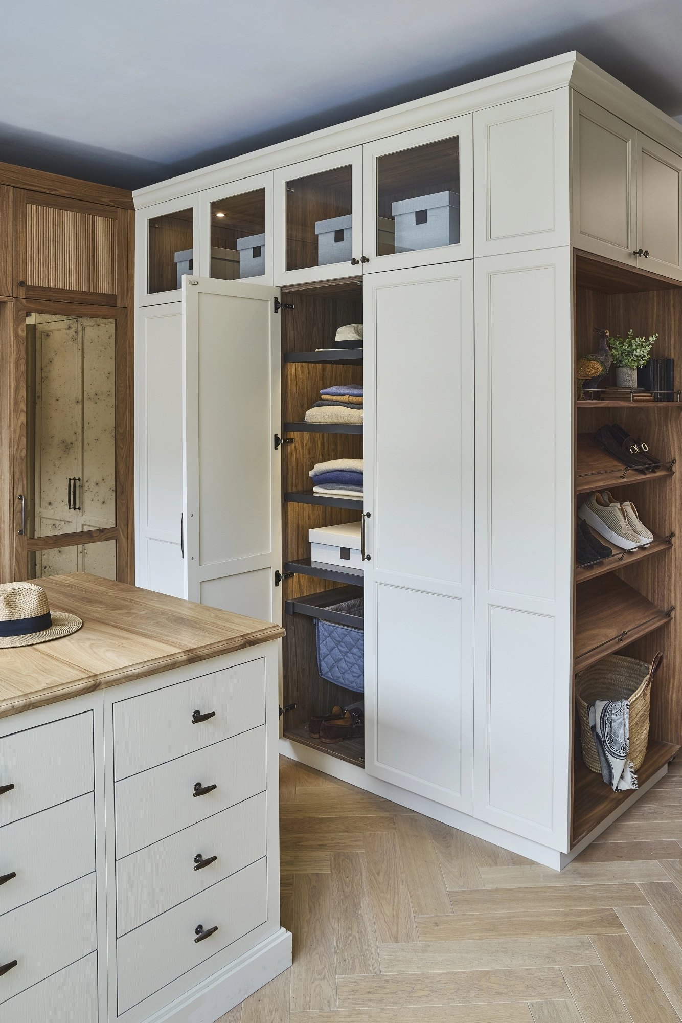 A modern walk-in closet with white cabinets, glass-paneled upper doors, wooden open shelves displaying shoes and baskets, a mirrored door, and a light wood island with drawers and a hat on top.