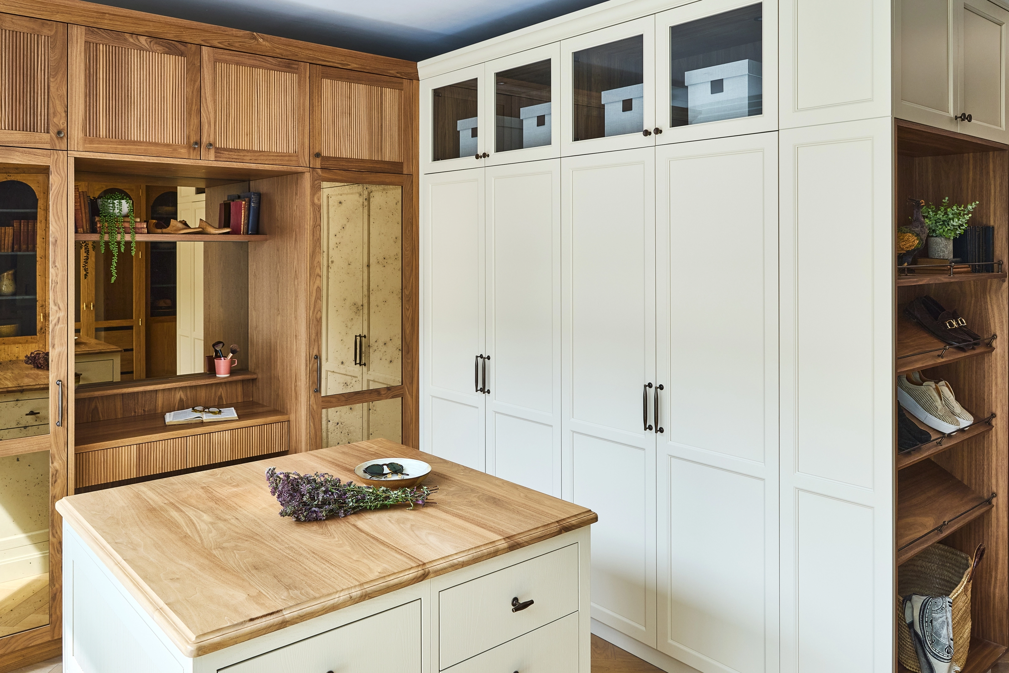 A modern walk-in closet with white cabinets, glass-paneled upper doors, wooden open shelves displaying shoes and baskets, a mirrored door, and a light wood island with drawers and a hat on top kitchen