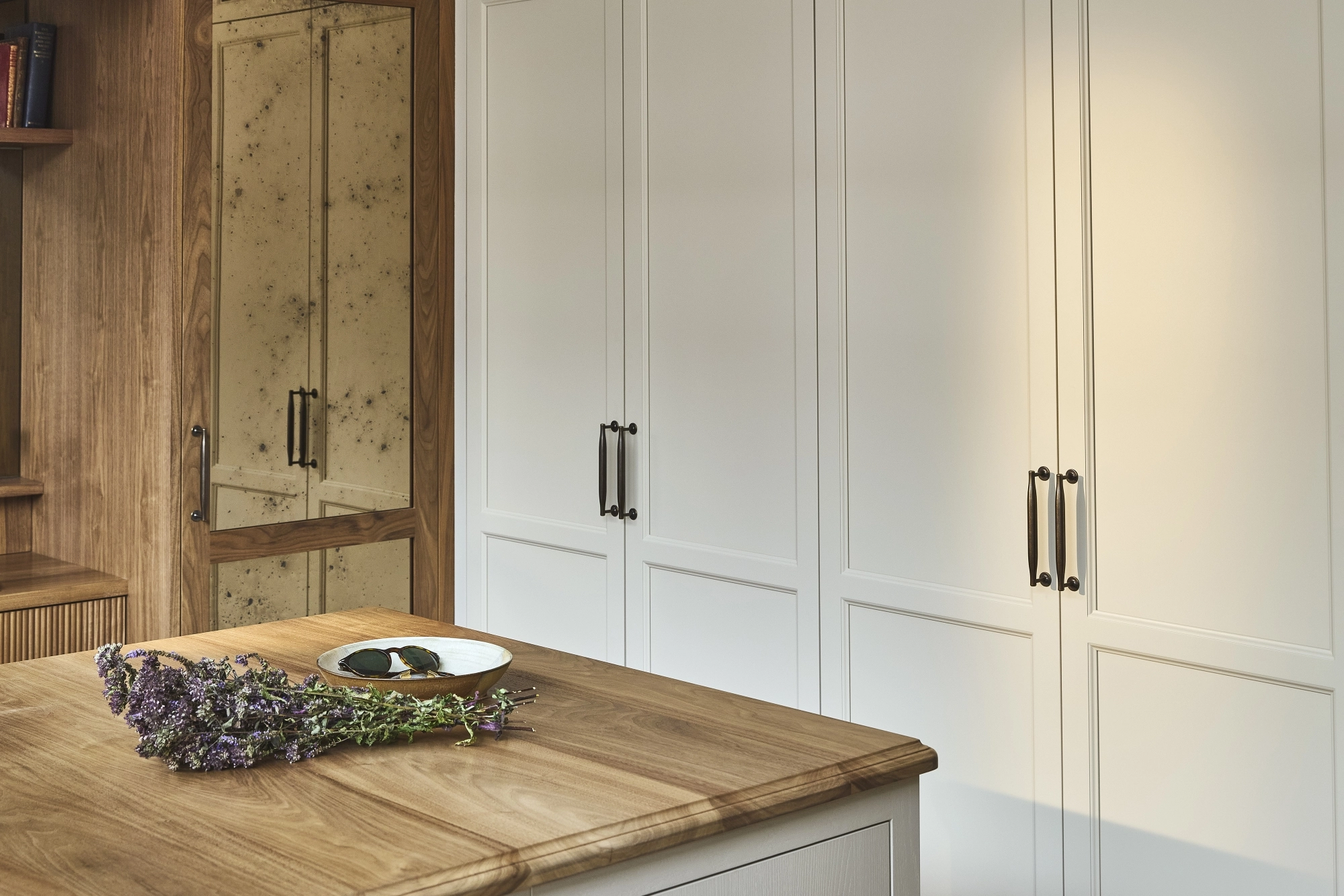A modern kitchen with white paneled cabinets, black handles, and wood accents. A kitchen island with a wood countertop holds a plate with two dark stones and a bundle of purple flowers.