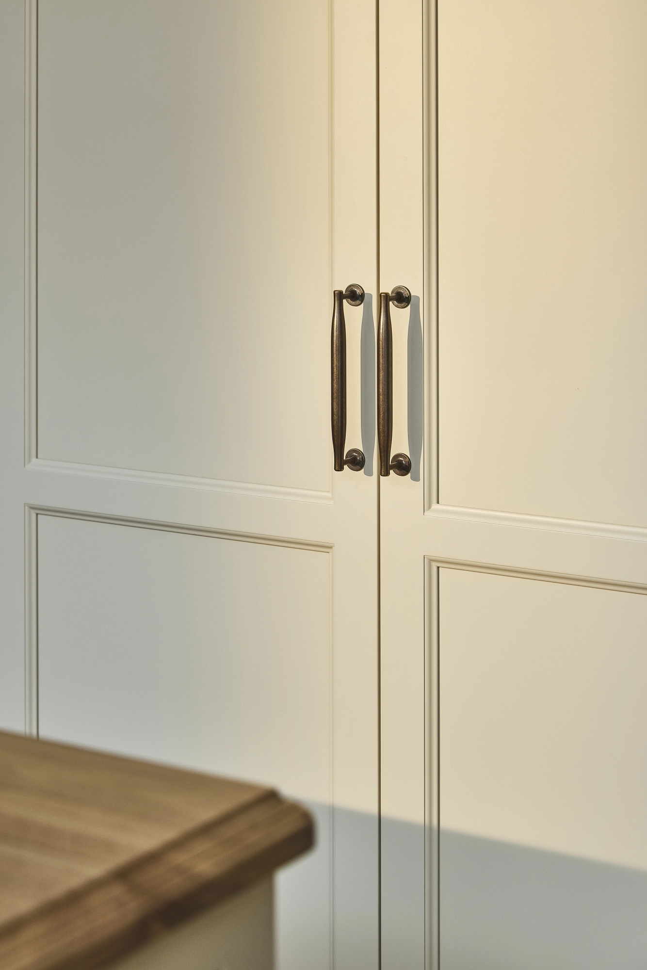 Cream-colored cabinet doors with dark metal handles, featuring a simple panel design. The edge of a wooden countertop is visible in the lower left corner of the image
