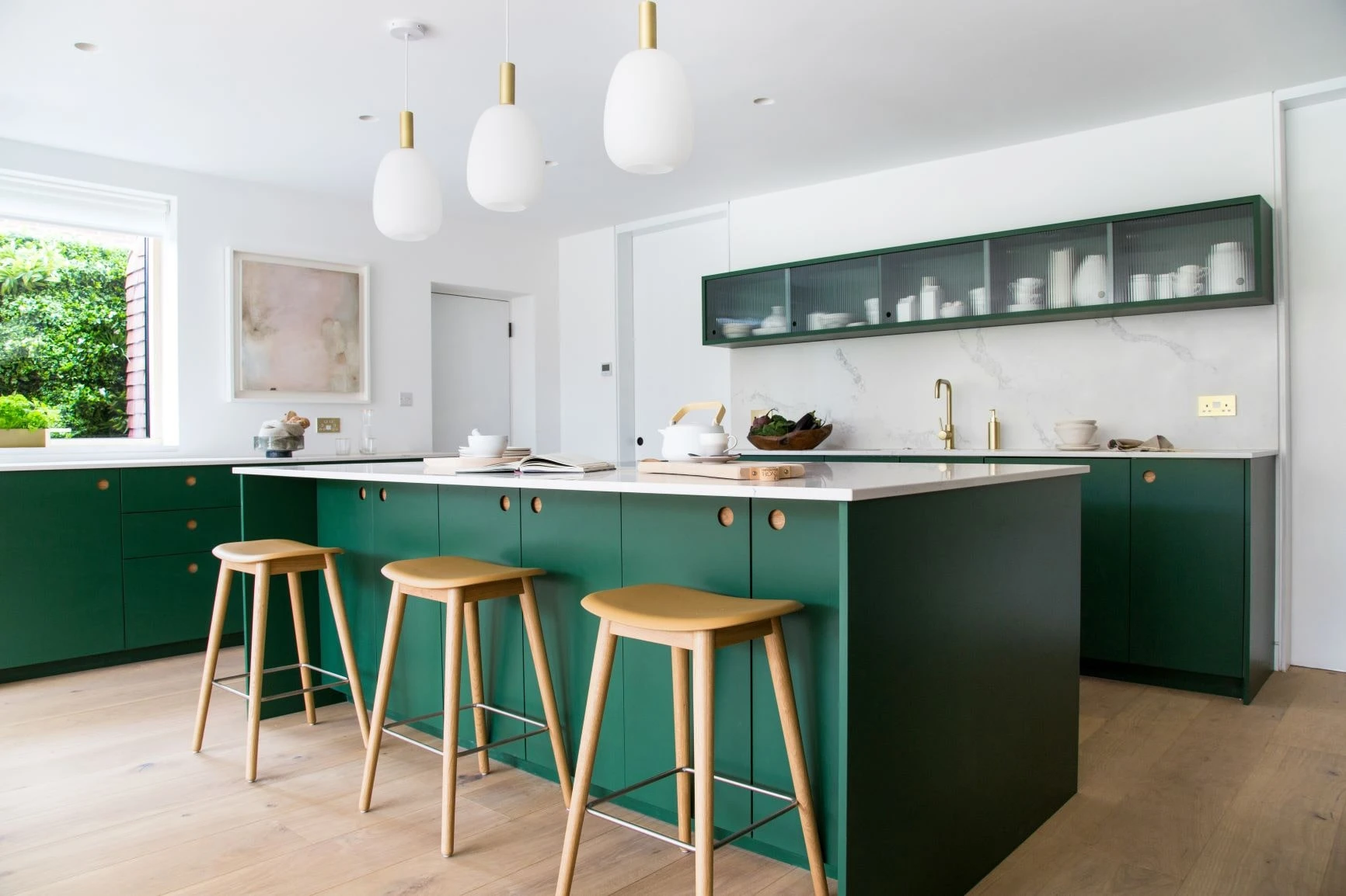 Wiveton Fern green Ladbroke doors blend seamlessly with oak accents and reeded glass cabinets, reflecting the surrounding greenery in this stunning minimalist kitchen. 