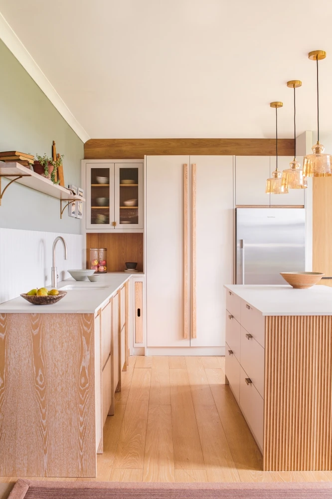 Modern kitchen with light wood cabinetry and white countertops. A large island features drawers and a wooden bowl. Pendant lights hang above. A fridge is integrated into floor-to-ceiling cabinets. Green and white walls add contrast.