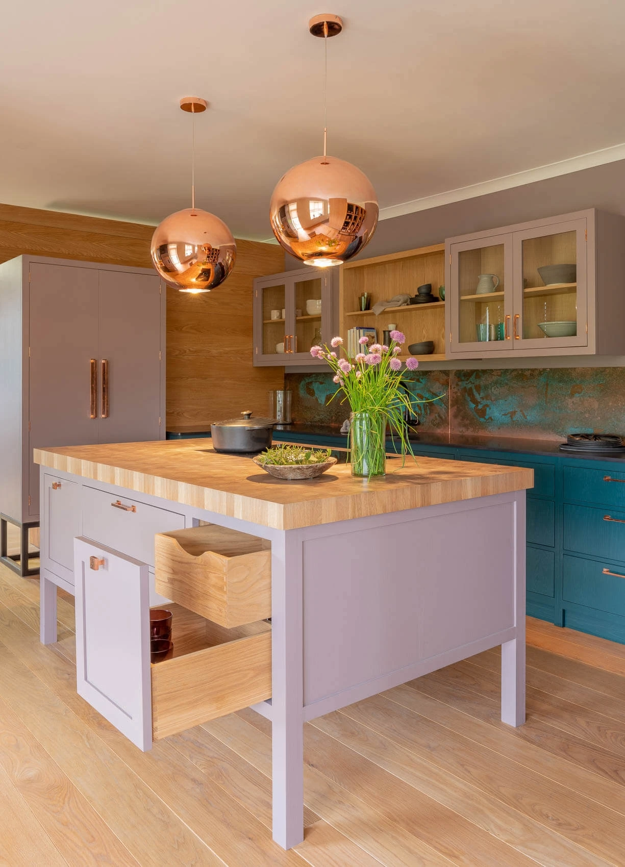 A modern kitchen with a wooden island featuring open drawers. Two copper pendant lights hang above. The countertop holds a vase of flowers and a bowl. Stainless steel appliances and glass-front cabinets are in the background.