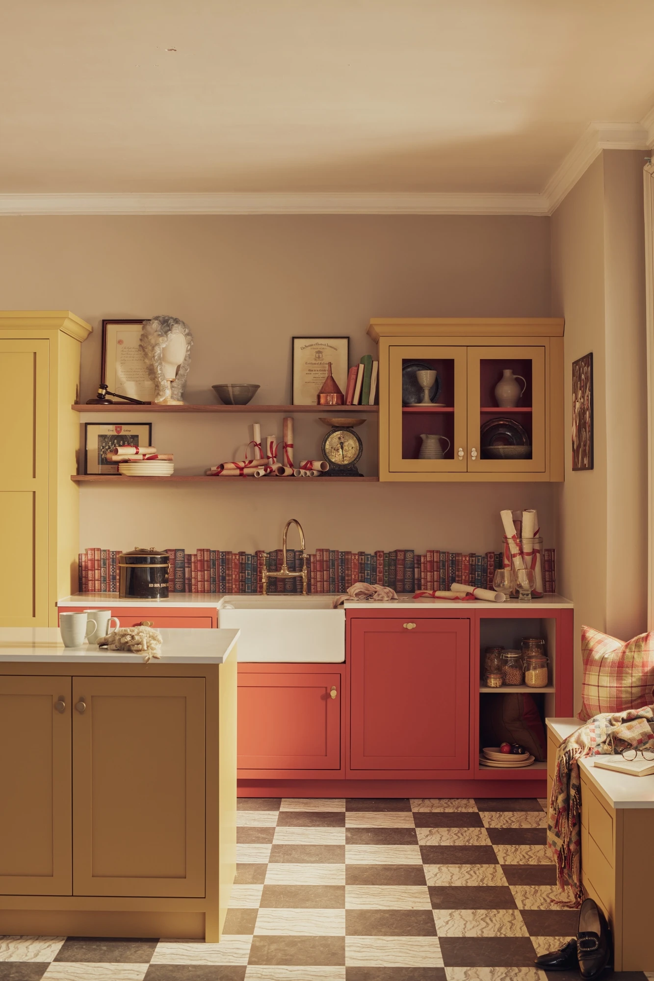 A cozy kitchen with red and yellow cabinets, a farmhouse sink, and checkered floor. Open shelves display books, a clock, and decorative items. Sunlight streams in, illuminating a mug on the counter and a cushioned bench