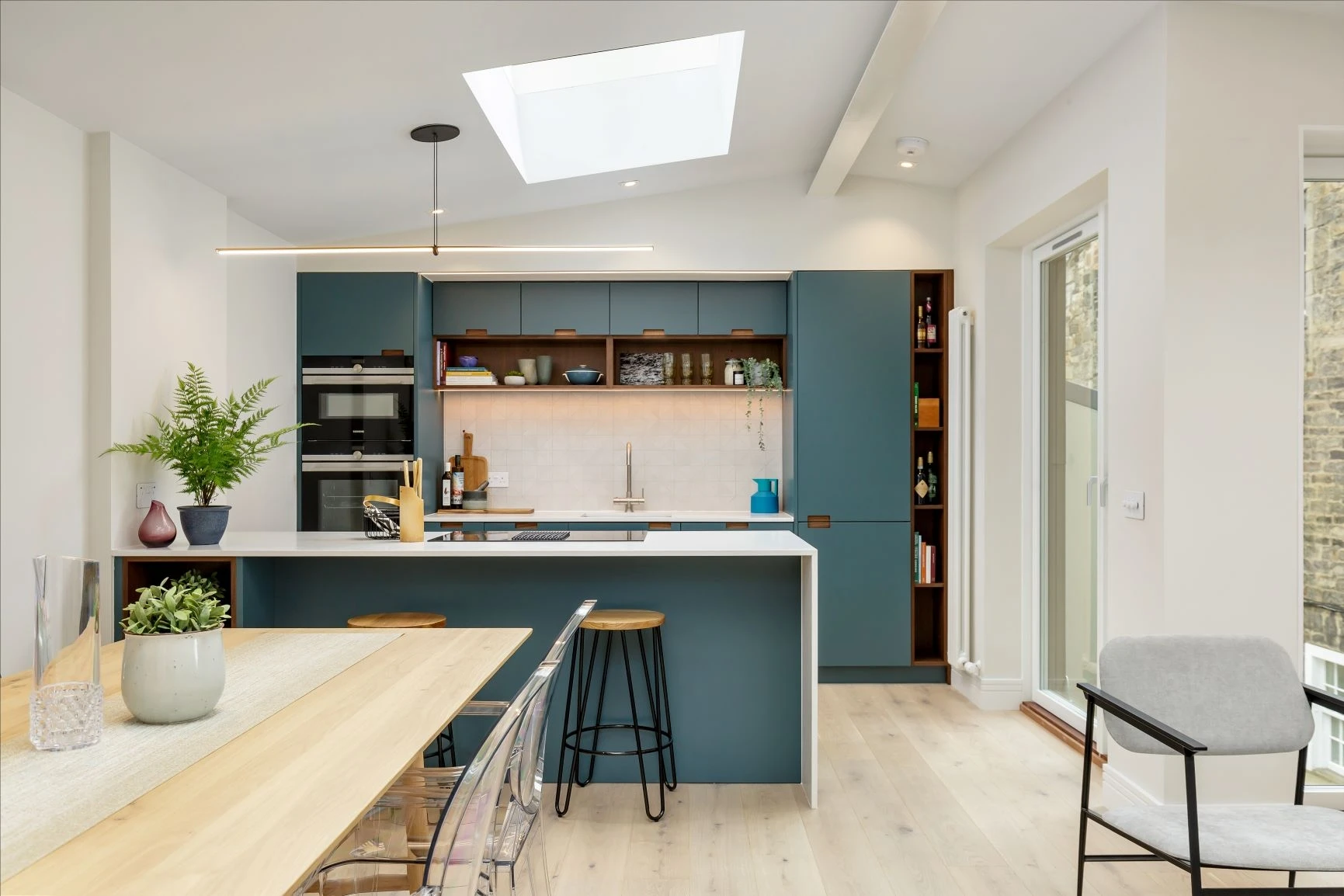 Modern kitchen interior with a skylight. Features teal cabinets, a white countertop island with two wooden stools, and built-in appliances. A wooden dining table with clear chairs and potted plants enhance the cozy atmosphere. Natural light fills the room.