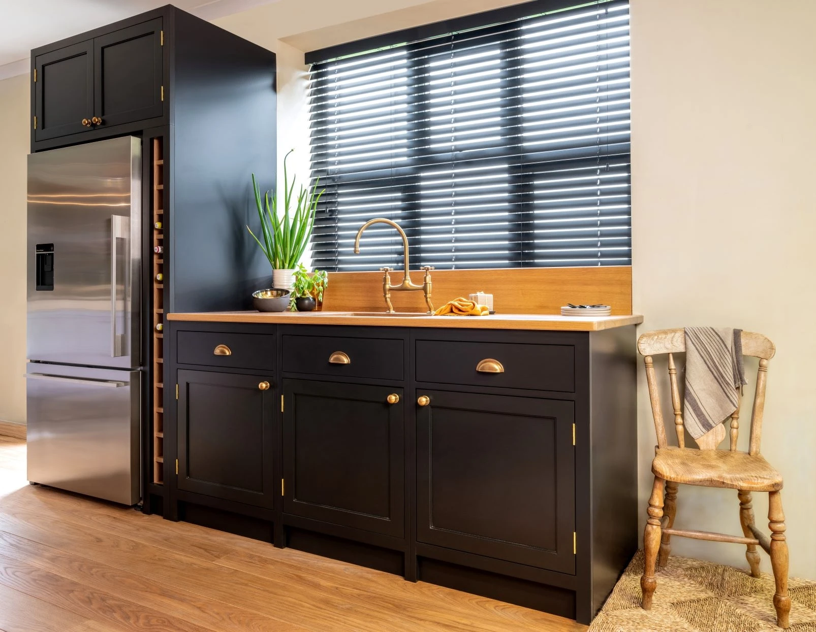 Modern kitchen with black cabinets, a stainless steel fridge, and a wooden countertop. A potted plant and dishes are on the counter. Large window with horizontal blinds behind the sink. A wooden chair with a towel is beside the cabinets on a wooden floor.
