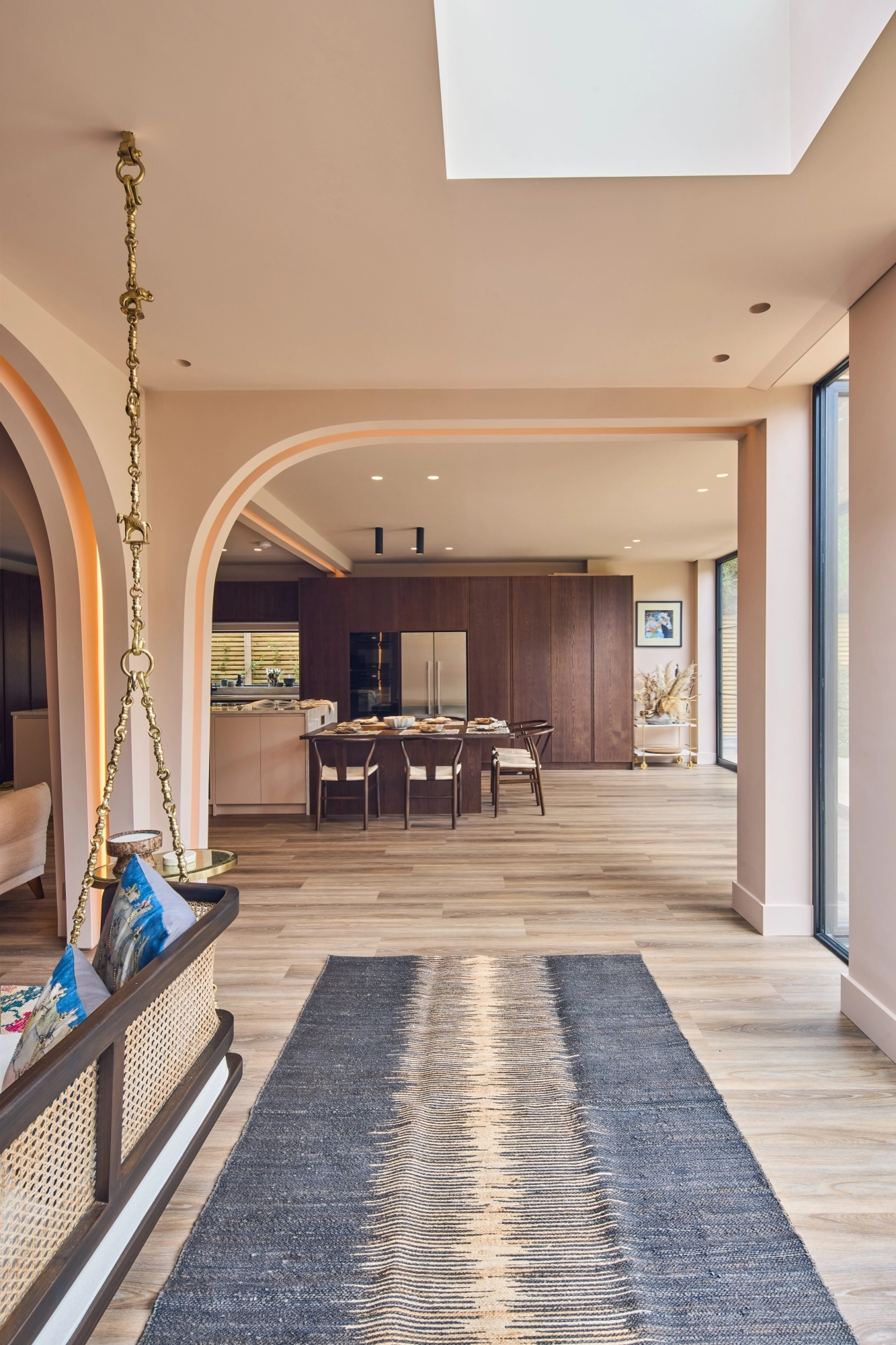 A cozy kitchen with a mix of styles. Light blue cabinets complement a marbled countertop and backsplash. A wooden shelf holds books and dishes, while a maroon island features a cutting board and red pot. Plants hang above the sink for decoration.