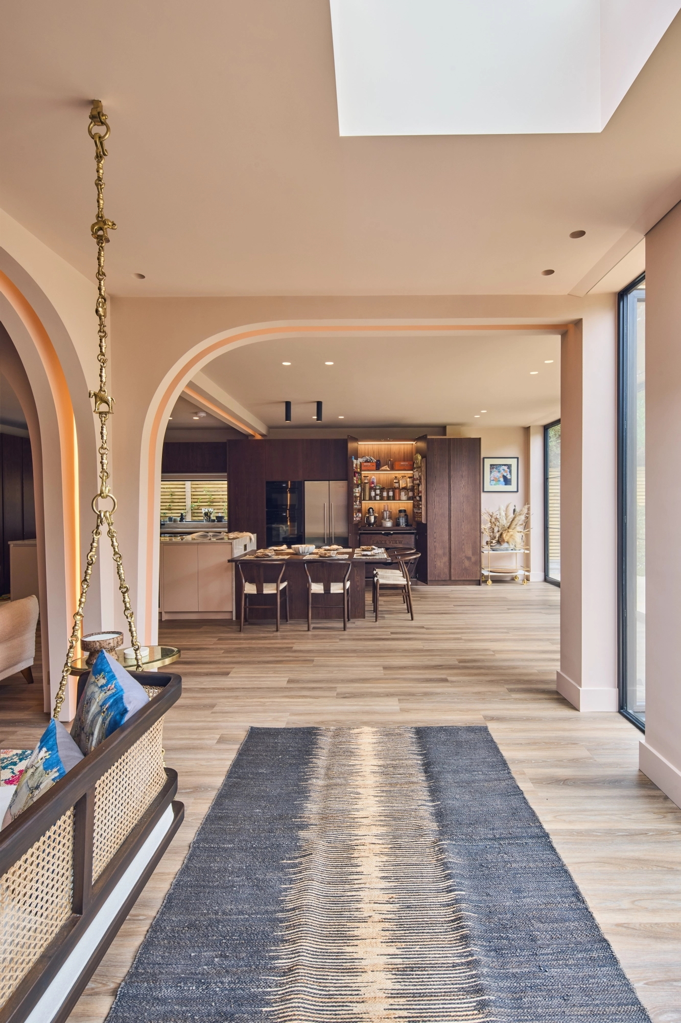 A cozy kitchen with a mix of styles. Light blue cabinets complement a marbled countertop and backsplash. A wooden shelf holds books and dishes, while a maroon island features a cutting board and red pot. Plants hang above the sink for decoration.