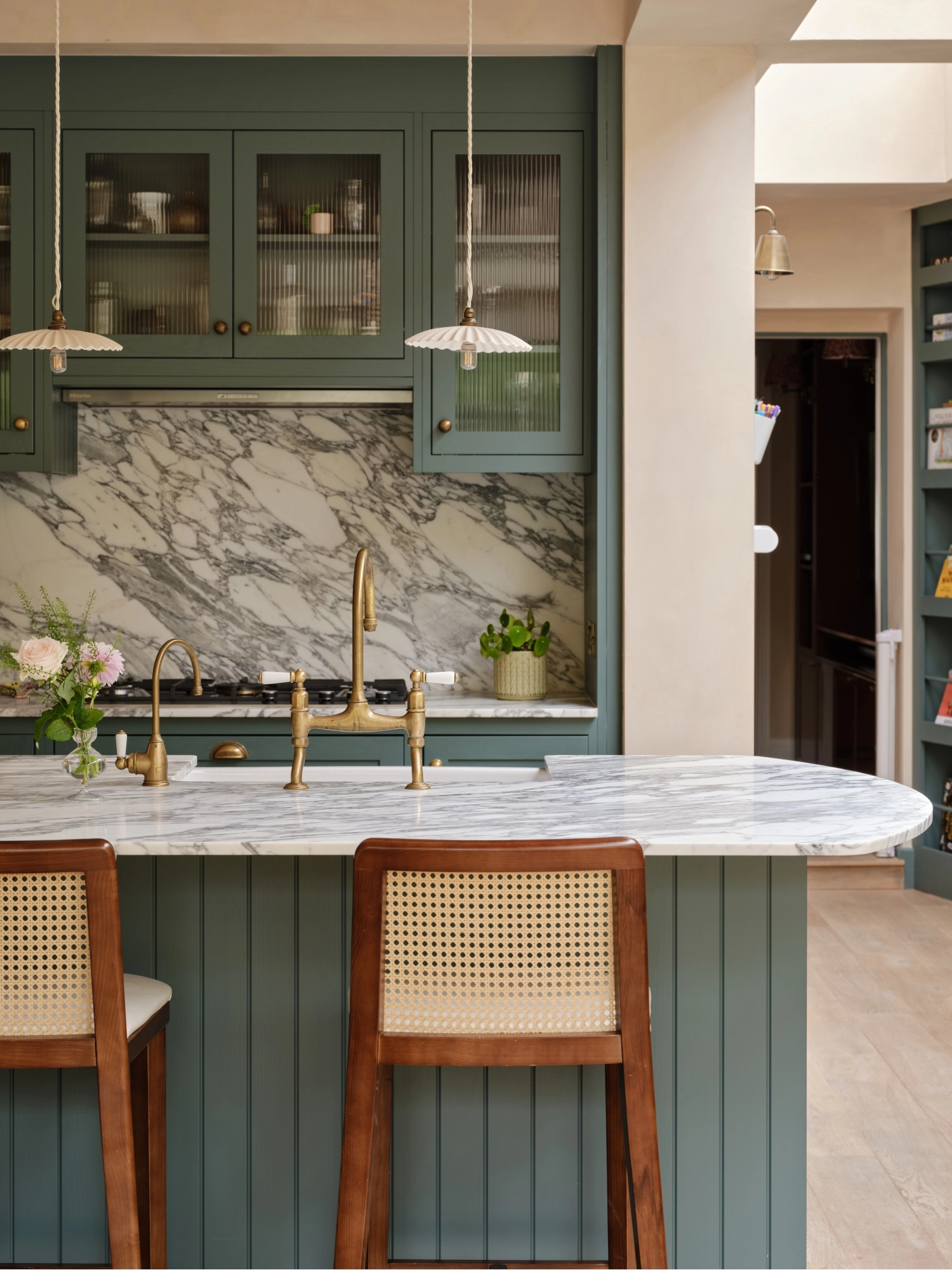 A cozy kitchen with a mix of styles. Light blue cabinets complement a marbled countertop and backsplash. A wooden shelf holds books and dishes, while a maroon island features a cutting board and red pot. Plants hang above the sink for decoration.