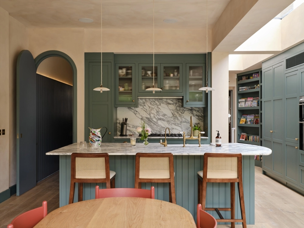 An elegant townhouse kitchen in custom green Shaker cabinetry with marble worktops, integrated seating and seamless garden connection.
