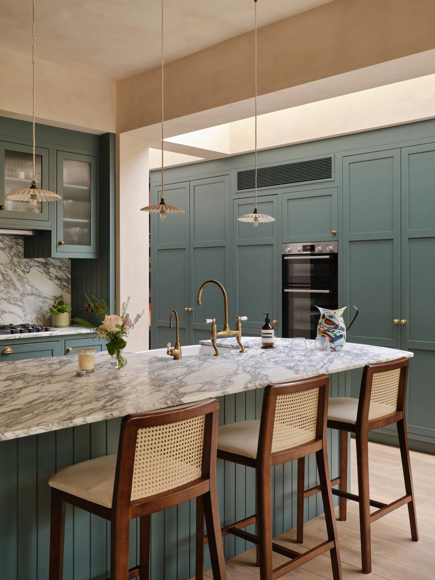 A cozy kitchen with a mix of styles. Light blue cabinets complement a marbled countertop and backsplash. A wooden shelf holds books and dishes, while a maroon island features a cutting board and red pot. Plants hang above the sink for decoration.
