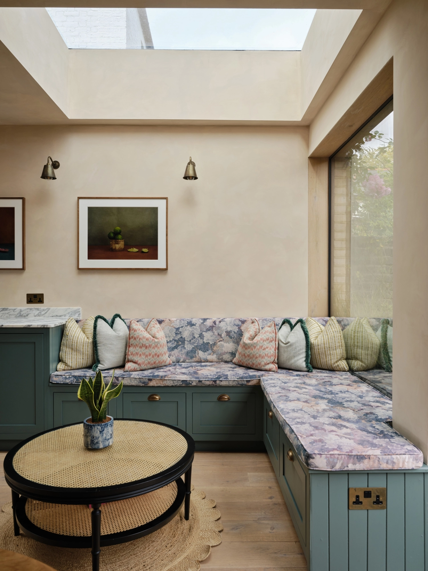 A cozy kitchen with a mix of styles. Light blue cabinets complement a marbled countertop and backsplash. A wooden shelf holds books and dishes, while a maroon island features a cutting board and red pot. Plants hang above the sink for decoration.
