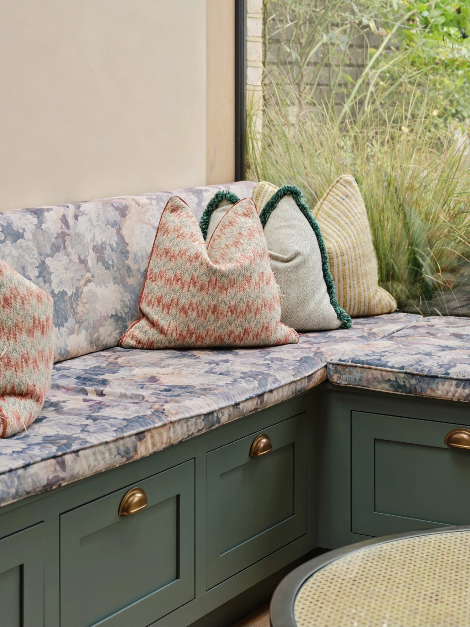 A cozy kitchen with a mix of styles. Light blue cabinets complement a marbled countertop and backsplash. A wooden shelf holds books and dishes, while a maroon island features a cutting board and red pot. Plants hang above the sink for decoration.