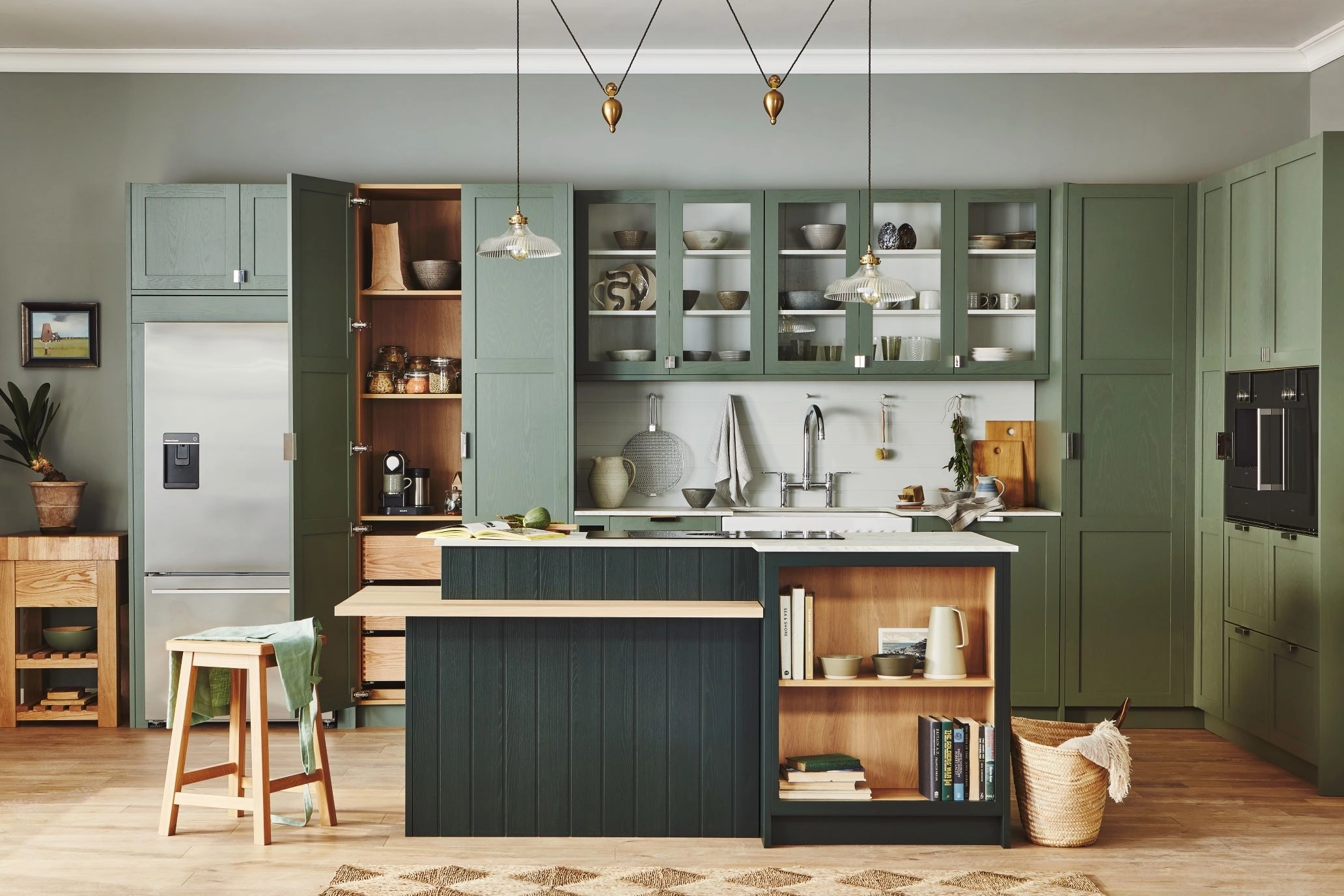 A modern kitchen with green cabinetry, light gray walls, and wooden accents. It features open shelves with dishes, a double-door fridge, a central island with a wooden stool, and pendant lights. Appliances include a built-in oven and coffee maker.