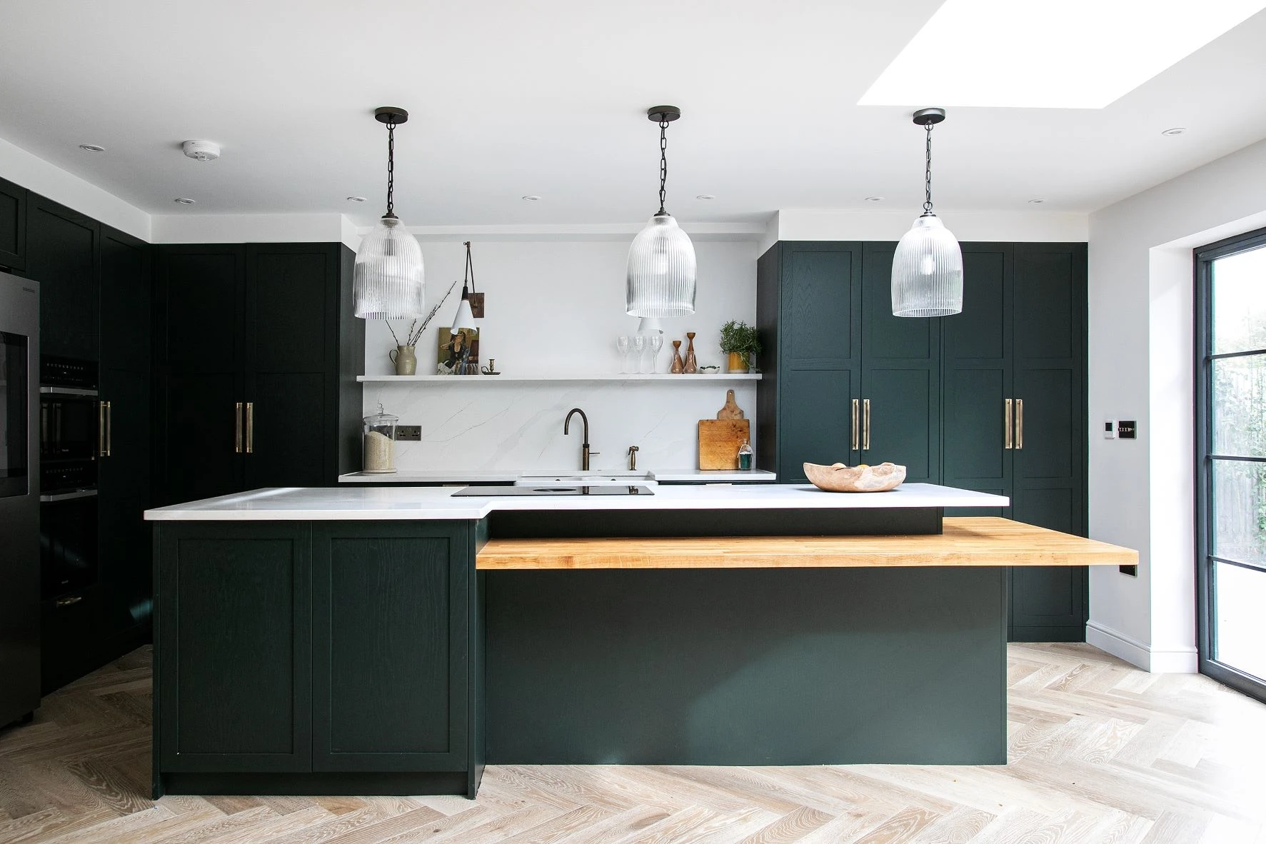 Modern kitchen with dark green cabinetry and a large island featuring a white countertop and wooden breakfast bar. Three pendant lights hang over the island. There are open shelves, marble backsplash, and stainless-steel appliances, with light herringbone flooring.