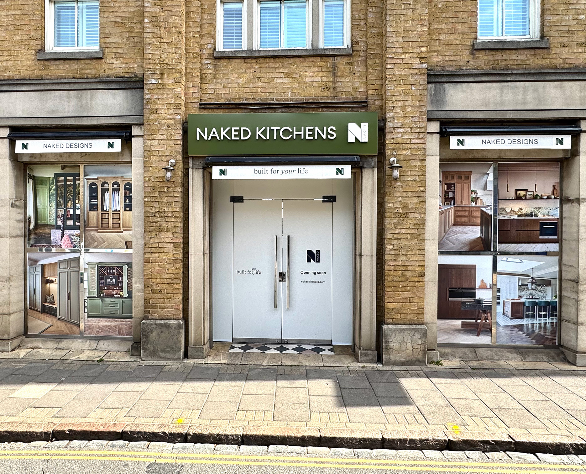 Street view of the Naked Kitchens showroom in a three-story yellow brick building. The central entrance features a green sign and white doors labeled Coming Soon. Flanking windows for Naked Designs and Naked Studio display various interior kitchen and furniture styles.